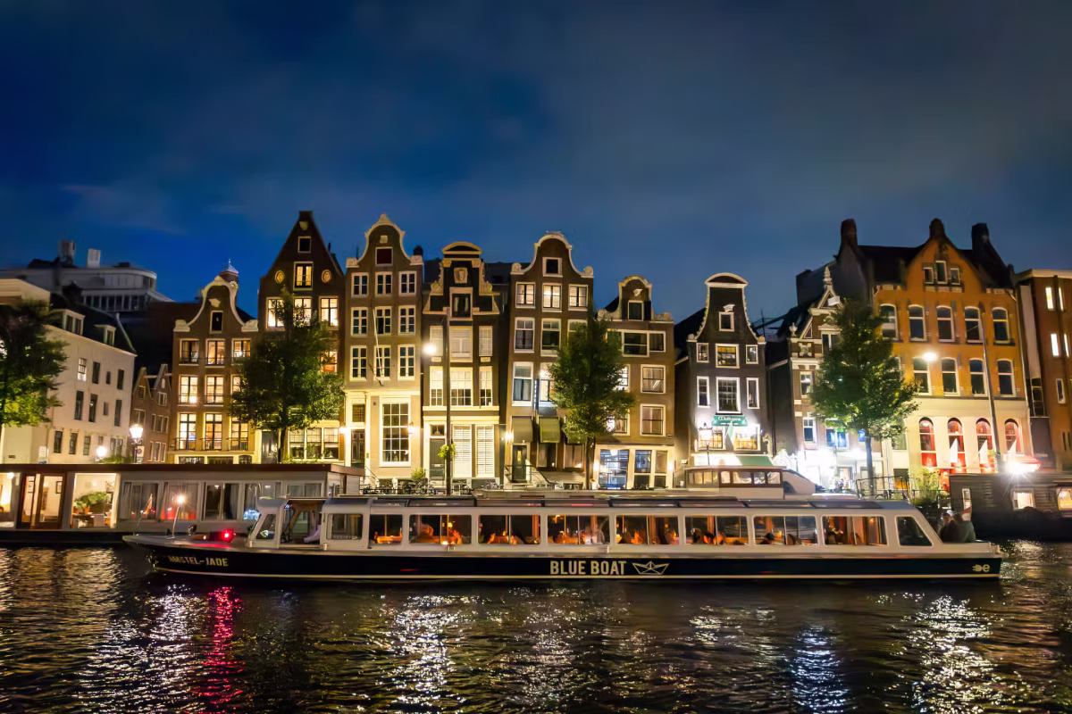 A canal cruise boat passes by illuminated Amsterdam canal houses at night.