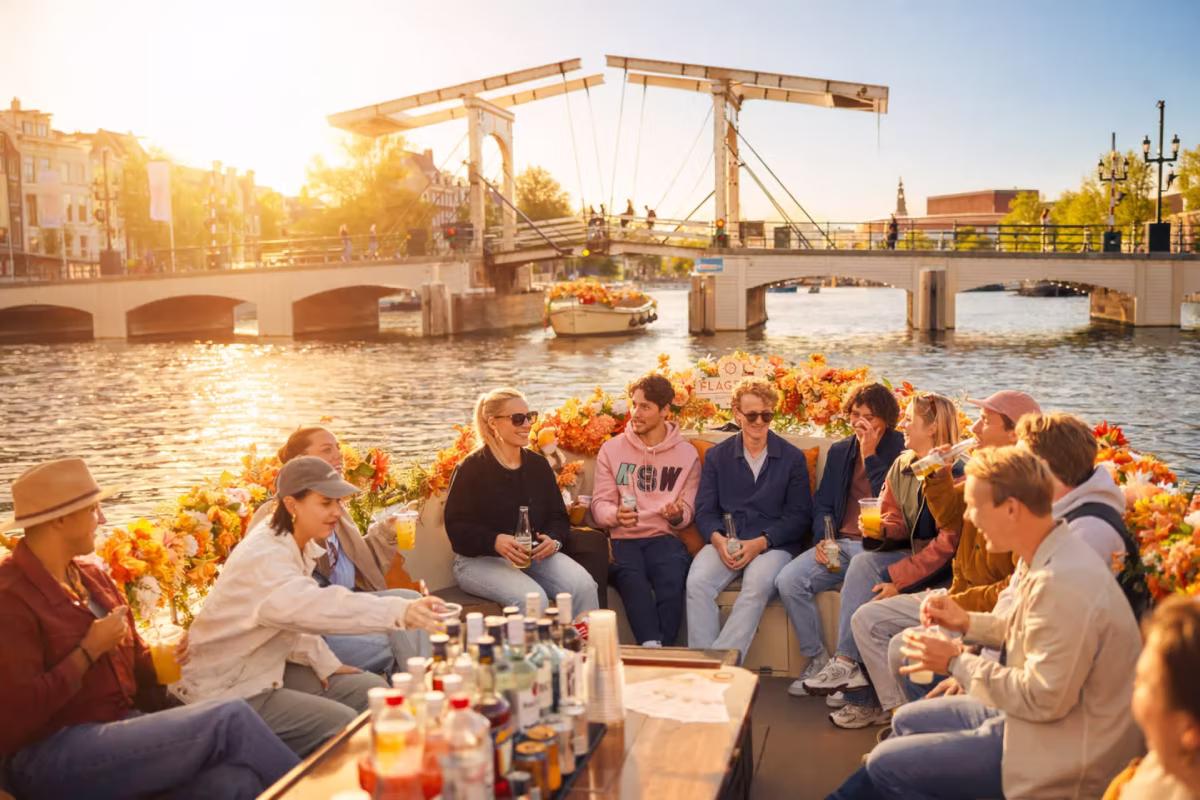A group of people enjoys drinks on an Amsterdam canal cruise at sunset near a bridge.